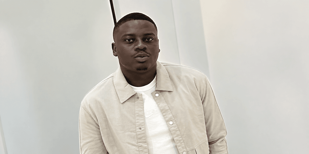 A young man in a light jacket poses against a simple backdrop