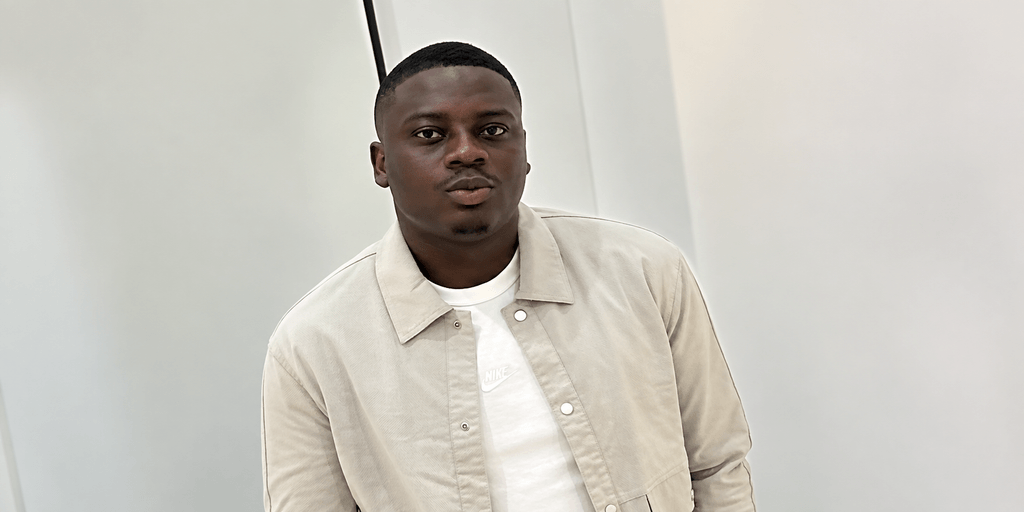 A young man in a light jacket poses against a simple backdrop