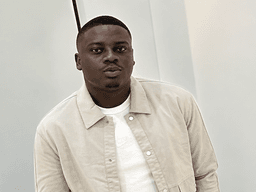 A young man in a light jacket poses against a simple backdrop