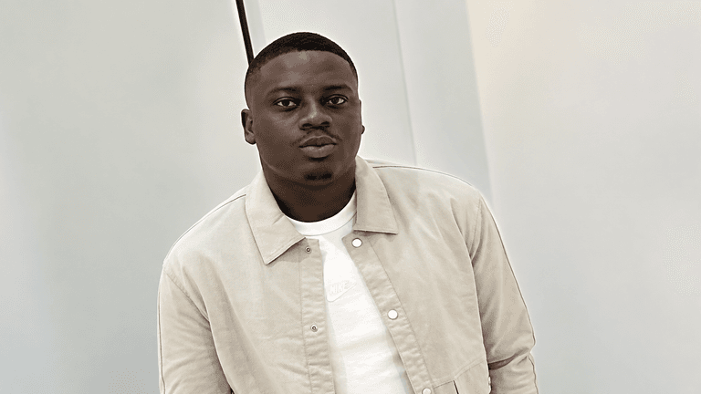 A young man in a light jacket poses against a simple backdrop