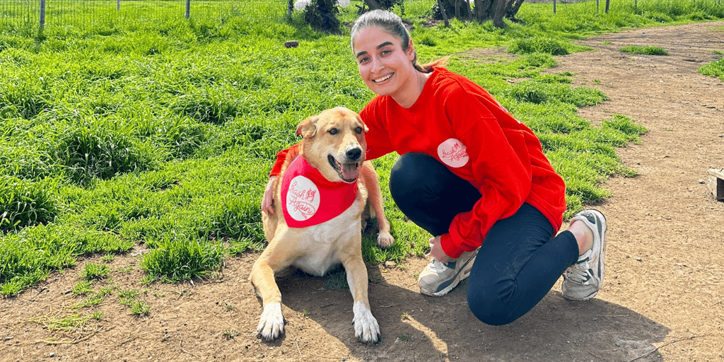 A person in a red sweater kneels next to a dog wearing a bandana