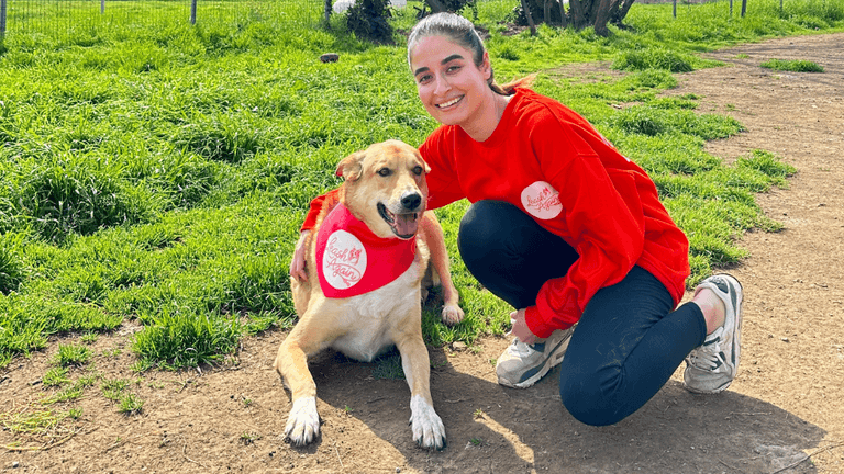 A person in a red sweater kneels next to a dog wearing a bandana