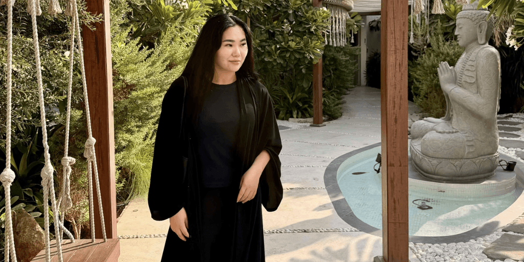 A woman in a black robe stands in a serene garden with a stone statue.