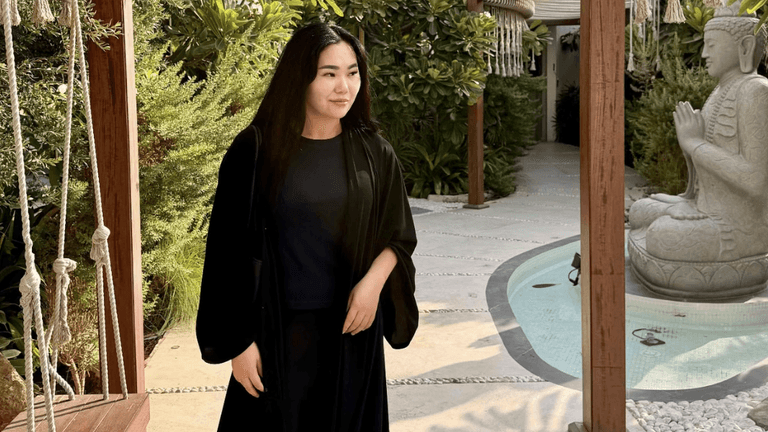 A woman in a black robe stands in a serene garden with a stone statue.