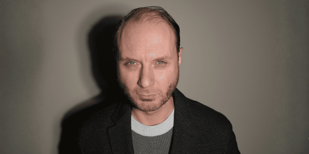 Man with short hair and beard, wearing a grey sweater and black jacket, serious expression