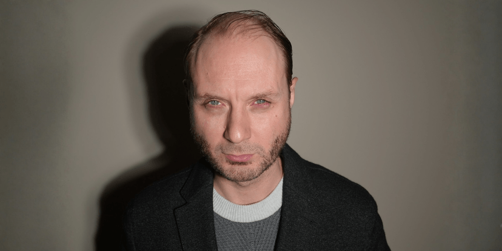 Man with short hair and beard, wearing a grey sweater and black jacket, serious expression
