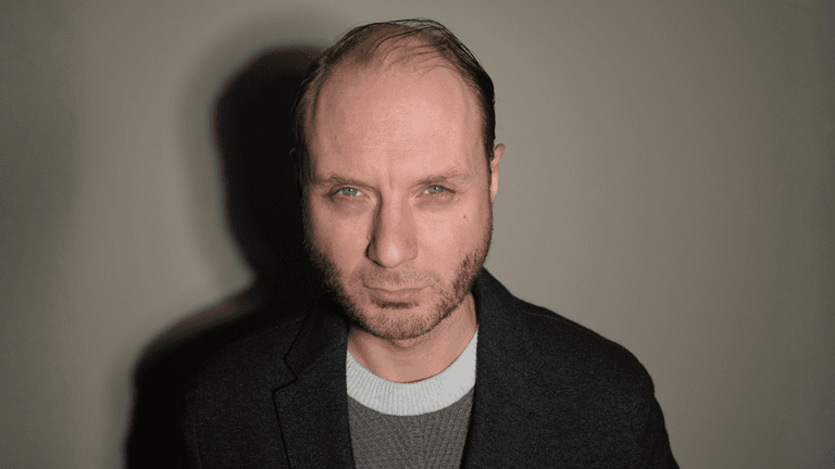 Man with short hair and beard, wearing a grey sweater and black jacket, serious expression