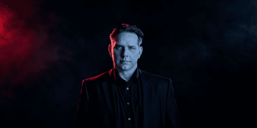 A man in a black suit stands against a dark, smoky background with red lighting