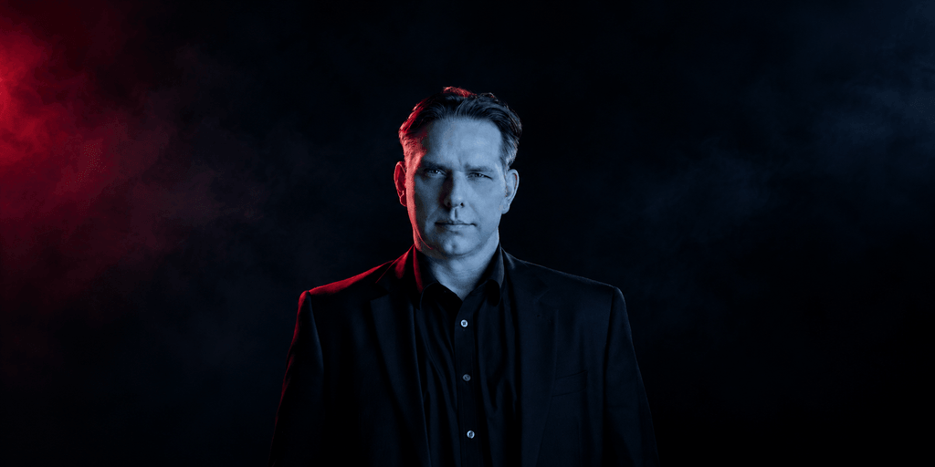 A man in a black suit stands against a dark, smoky background with red lighting