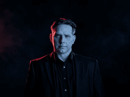 A man in a black suit stands against a dark, smoky background with red lighting