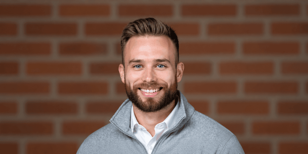 Smiling man with a beard wearing a gray sweater against a brick background