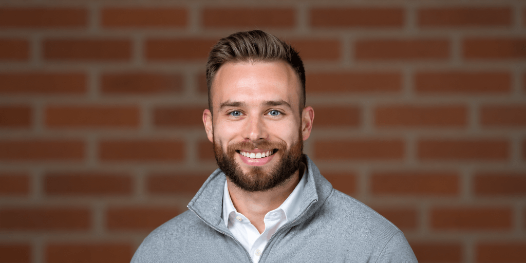 Smiling man with a beard wearing a gray sweater against a brick background