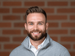 Smiling man with a beard wearing a gray sweater against a brick background