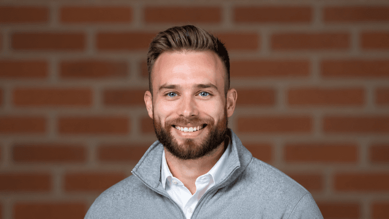 Smiling man with a beard wearing a gray sweater against a brick background