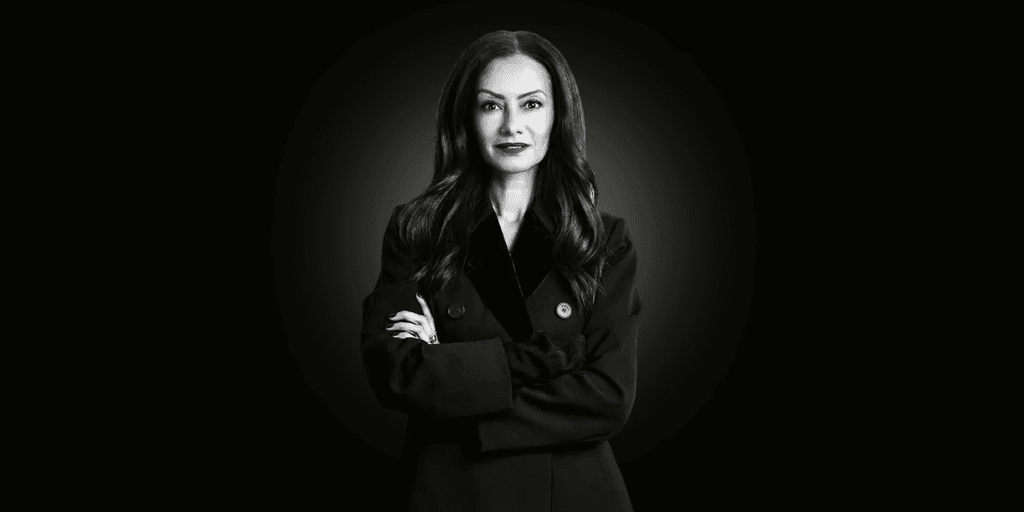A woman in a dark coat stands confidently with her arms crossed against a black background