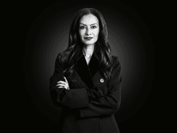 A woman in a dark coat stands confidently with her arms crossed against a black background