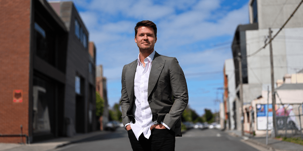 A man in a suit stands confidently on a city street with buildings nearby