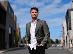 A man in a suit stands confidently on a city street with buildings nearby