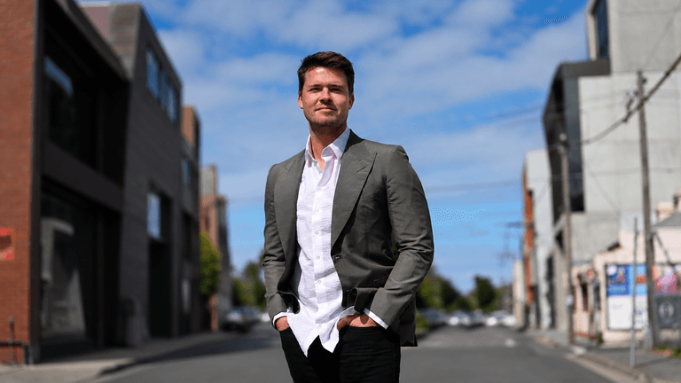 A man in a suit stands confidently on a city street with buildings nearby