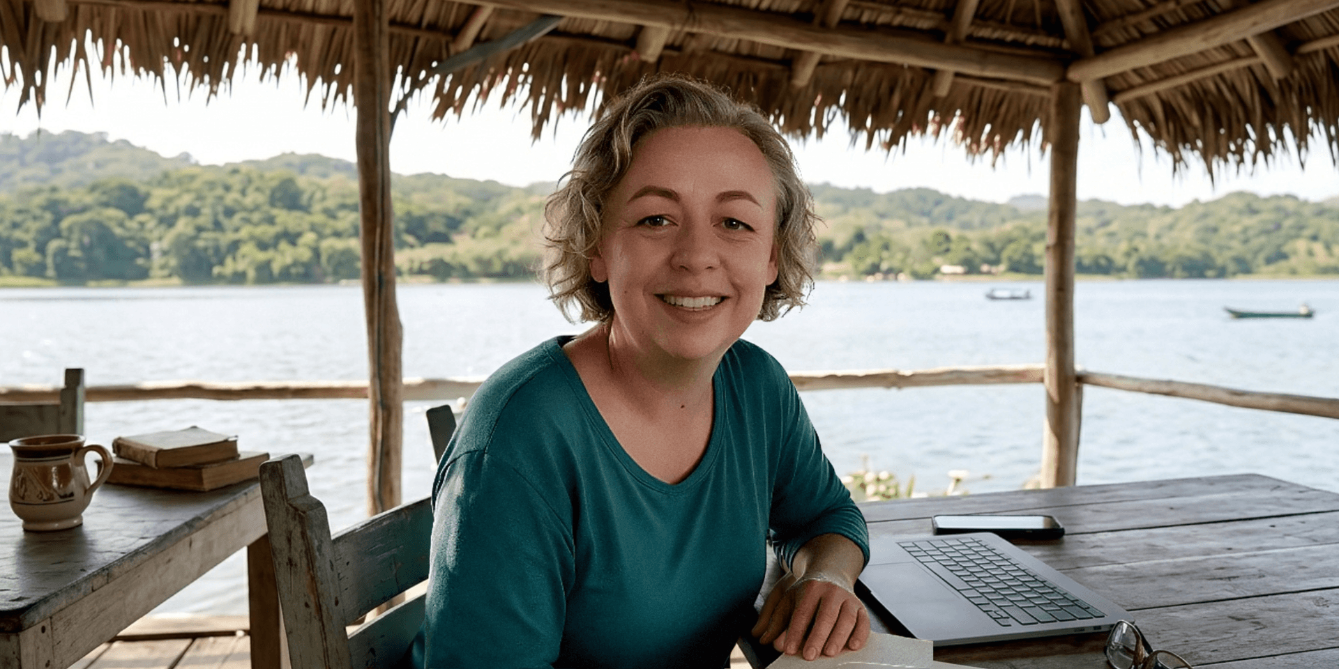 Smiling woman sitting at a wooden table by a lake, with a laptop and books.