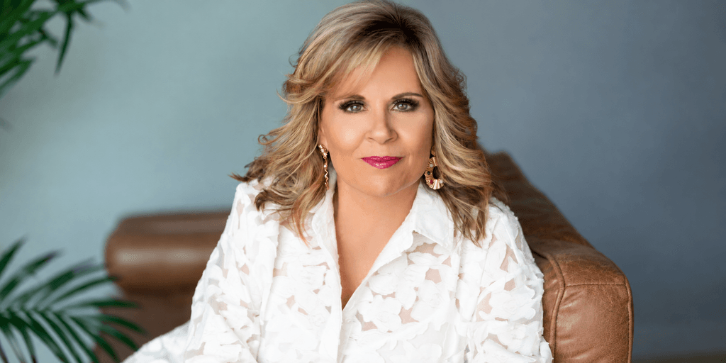 A woman with blonde hair in a white floral blouse sitting on a couch