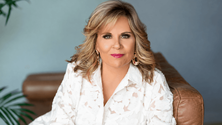 A woman with blonde hair in a white floral blouse sitting on a couch