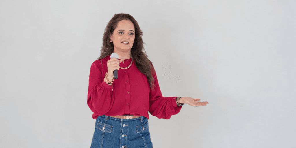 A woman in a red blouse holds a microphone while speaking or presenting