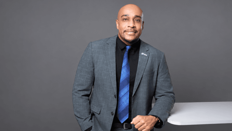 Man in a suit stands confidently beside a white table against a gray background