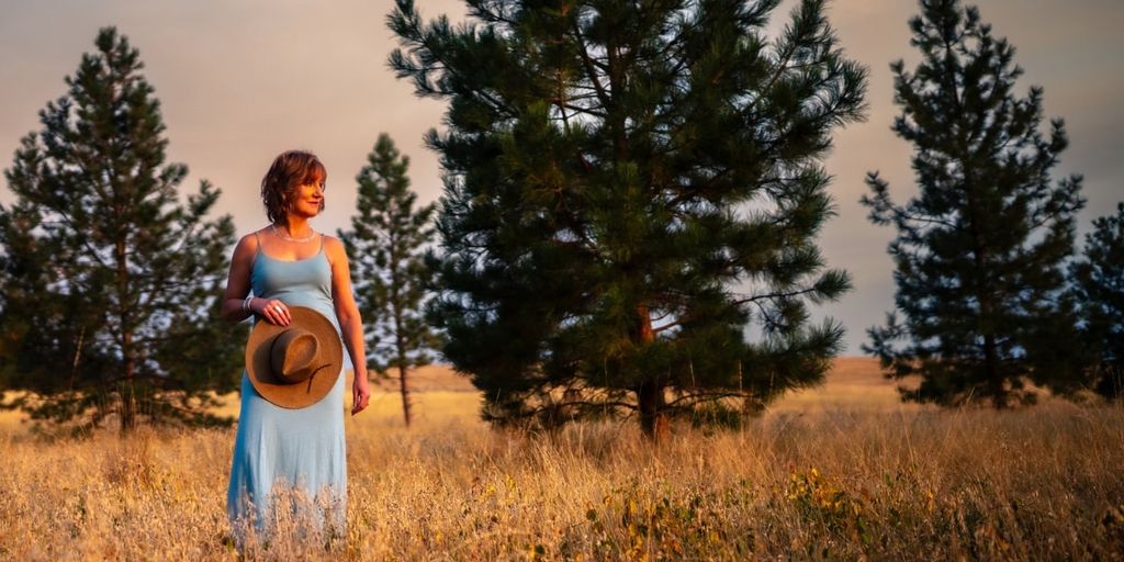 A woman in a light blue dress standing in a field, holding a hat, with pine trees in the background.