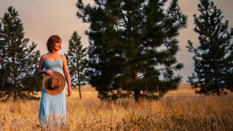 A woman in a light blue dress standing in a field, holding a hat, with pine trees in the background.