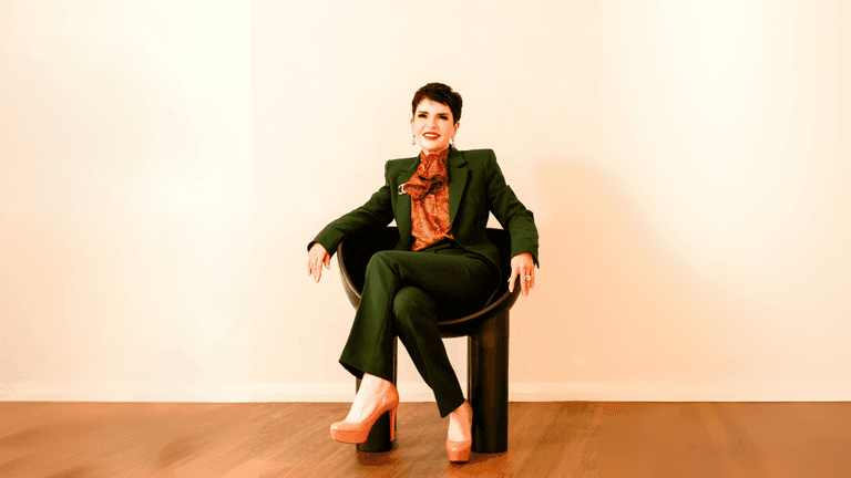 Portrait of a confident woman in a green suit, sitting in a modern chair with high heels, looking relaxed.