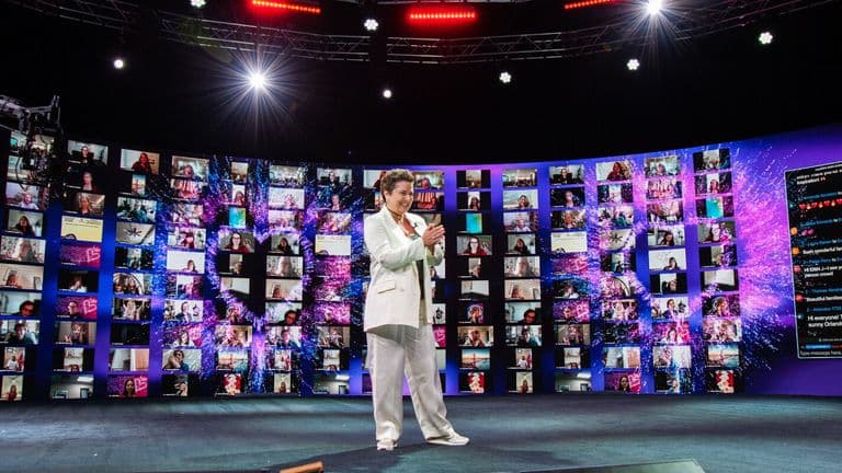 Josselyne Herman-Saccio speaking at an event, surrounded by virtual attendees displayed on large screens, celebrating her Evergreen Award.