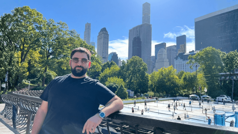 Alex Maghakian Gal leaning on railing in Central Park with Manhattan skyline behind.