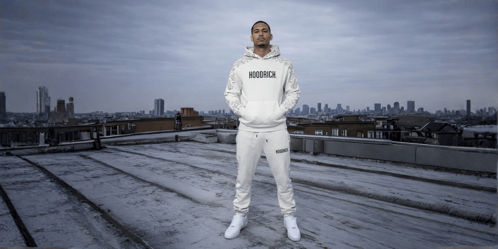 Model standing confidently in white hoodie and pants, overlooking city skyline on a rooftop.