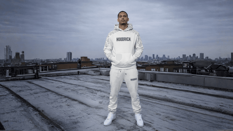 Model standing confidently in white hoodie and pants, overlooking city skyline on a rooftop.