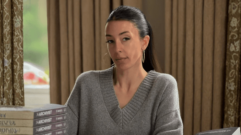 A woman in a gray sweater with hoop earrings, sitting at a table with copies of 'The Archivist' by Ti Mikkel, looking thoughtfully at the camera.