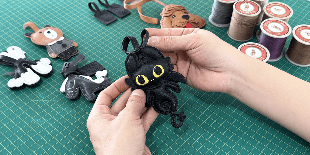 Hands holding a handcrafted black leather dragon charm on a workshop cutting mat.