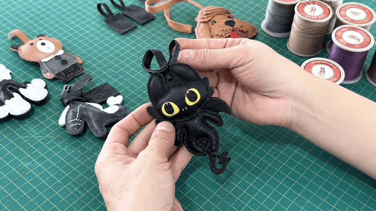 Hands holding a handcrafted black leather dragon charm on a workshop cutting mat.