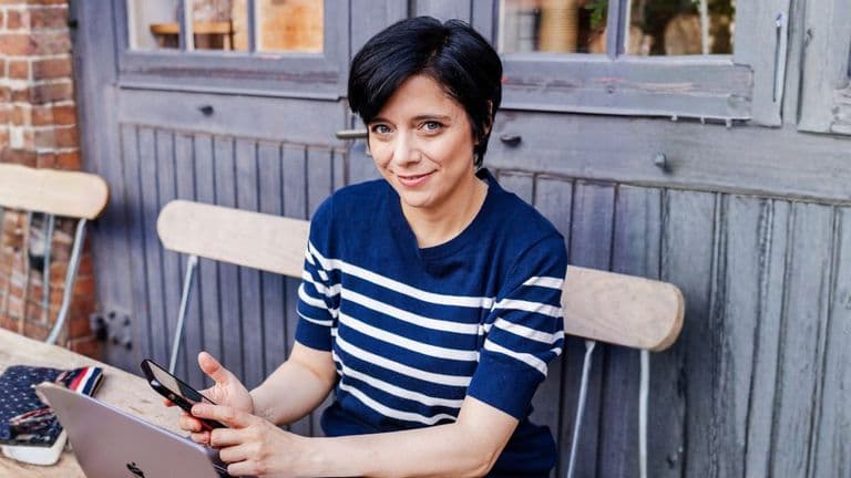 Woman using laptop and phone at outdoor café table, smiling confidently.