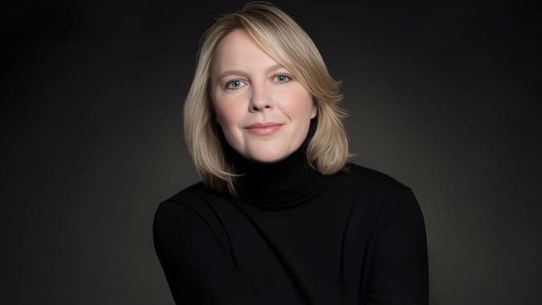 A person with shoulder-length blonde hair in a black turtleneck smiles gently against a dark background, conveying a calm and confident tone.