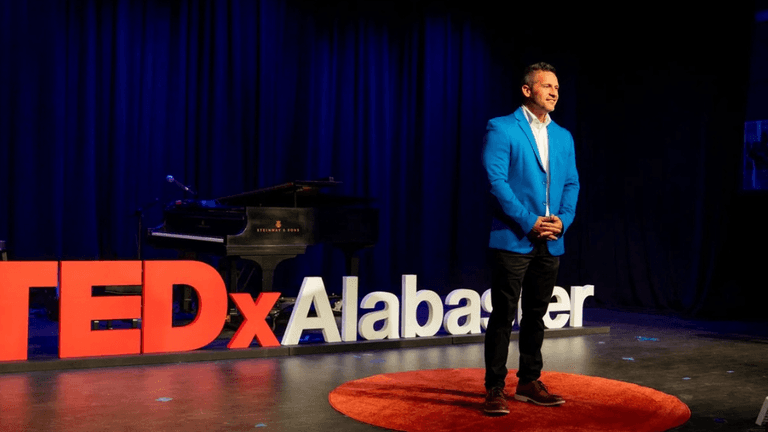 A man in a blue blazer stands on stage at TEDxAlabaster, with a piano and TEDx sign in the background.