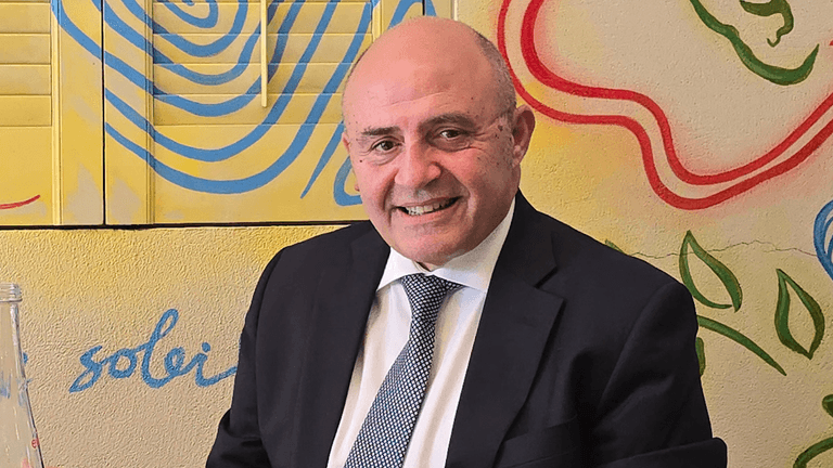 "Man seated at a table with a colorful mural in the background, wearing a dark suit and a patterned tie. He smiles warmly, exuding a relaxed and approachable demeanor."