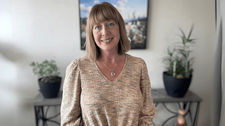 "A woman standing in front of a plant-filled table, smiling warmly. She wears a textured beige top and a pendant necklace. The background has a scenic photograph of nature."