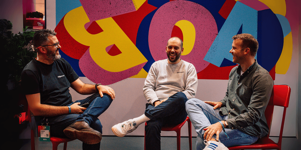 Three men sitting and laughing together in front of a colorful background at a business event, discussing ideas.