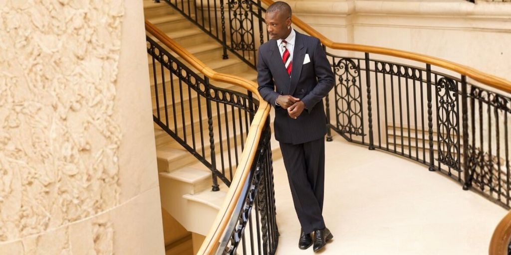 A man in a dark suit, white shirt, and red striped tie stands on a grand staircase with ornate railings, exuding confidence and elegance.
