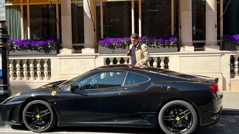 A man stands next to a black Ferrari sports car in London, showcasing elegance and professionalism in a city setting.