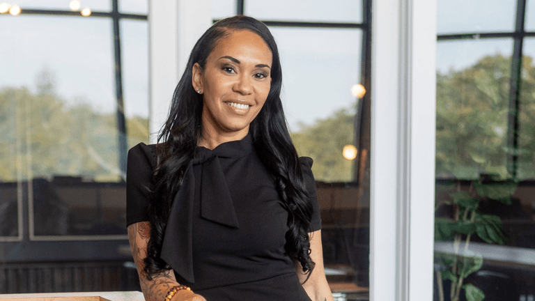 A woman in a black dress smiling confidently, standing in a bright, modern office with large windows and greenery in the background.