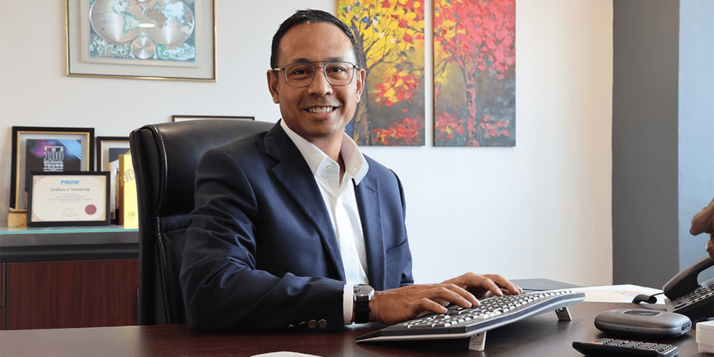 "A professional smiling man in a suit sitting at his desk in an office with framed certificates and artwork."