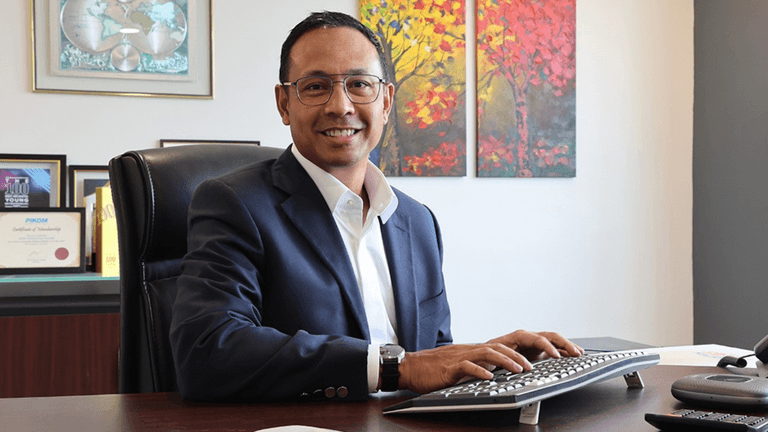 "A professional smiling man in a suit sitting at his desk in an office with framed certificates and artwork."