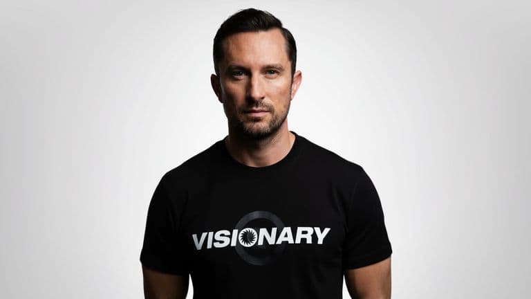 A man in a black "VISIONARY" t-shirt standing against a neutral background.